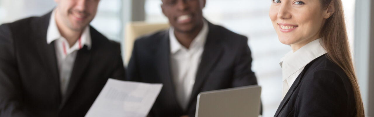 Attractive young businesswoman smiling for camera sitting at desk, young executive manager holding business meeting for black and white subordinates clients at background, female team lead portrait