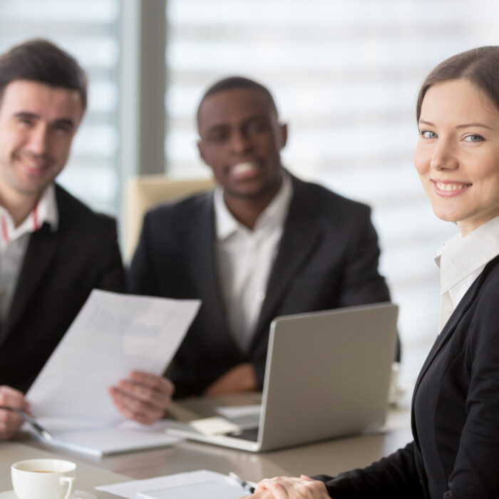 Attractive young businesswoman smiling for camera sitting at desk, young executive manager holding business meeting for black and white subordinates clients at background, female team lead portrait