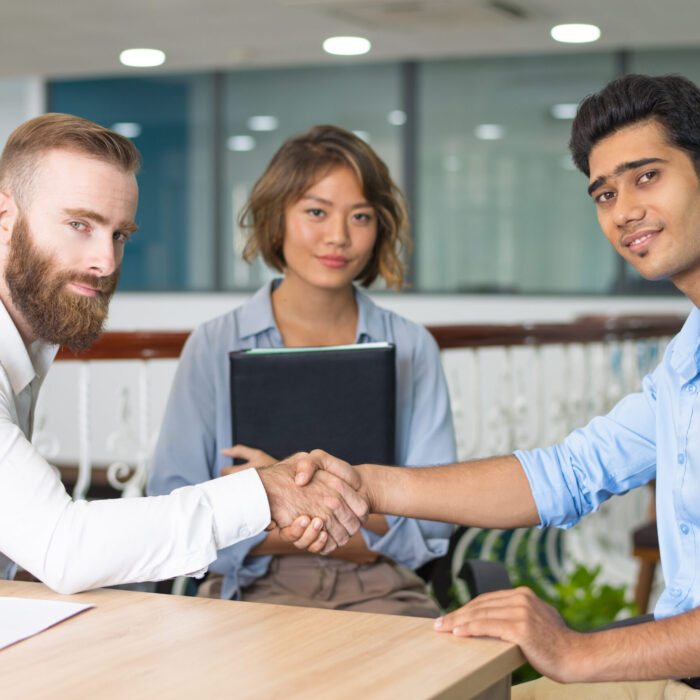 Content Indian candidate shaking hands with company leader at and of job interview. Asian HR manager posing in background. Job interview concept